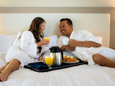 A couple in white bathrobes is relaxing on a bed, enjoying breakfast with orange juice, coffee, and other food items on a tray in front of them.