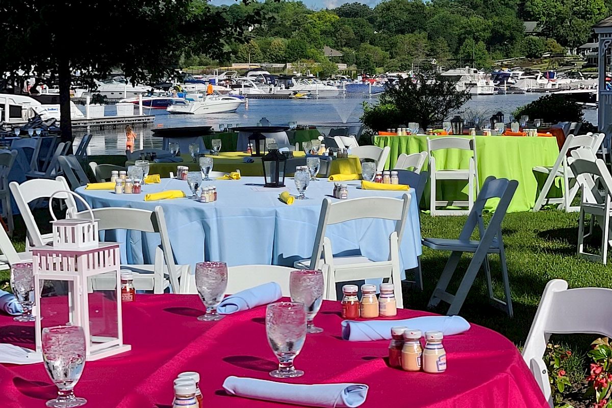 Outdoor tables set with colorful tablecloths, glassware, and lantern centerpieces by a waterfront area with boats in the background.
