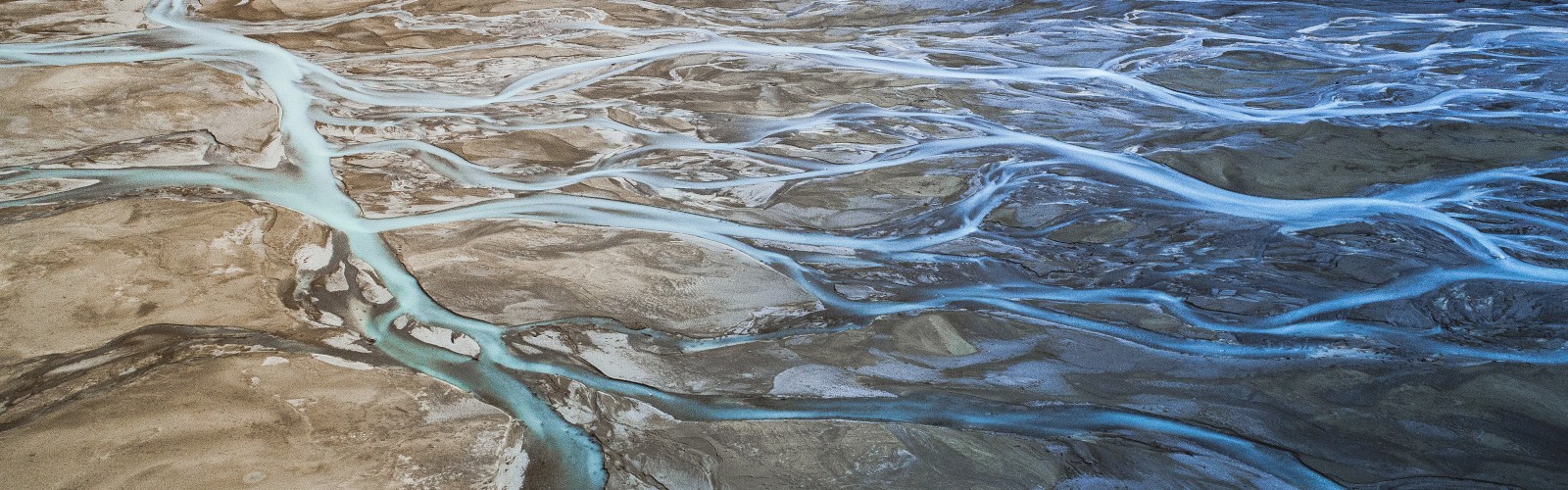 An aerial view showing a braided river flowing through a barren, rocky landscape with multiple interweaving channels of water in various shades of blue.