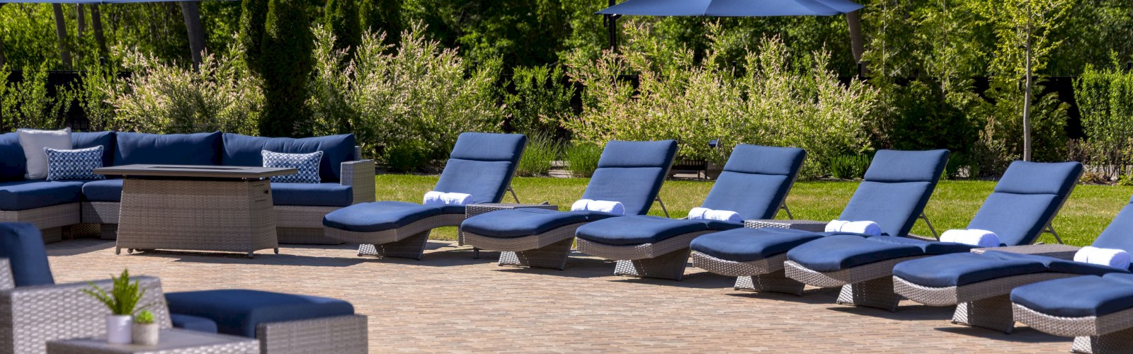 The image shows an outdoor patio area with blue lounge chairs, umbrellas, and a seating area. It's surrounded by trees and greenery.
