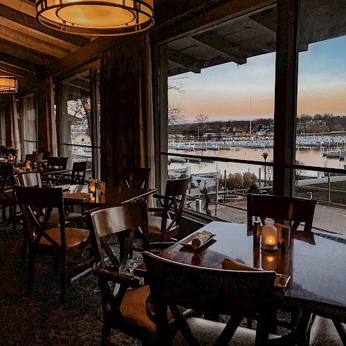 A chic, dimly lit restaurant interior with wooden chairs and tables, large windows overlooking a marina at sunset. End.
