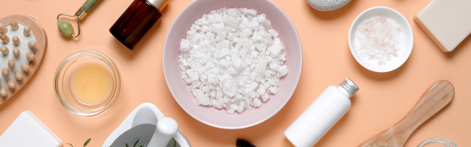 Various skincare items including oils, serums, soap, a brush, and a jade roller arranged around a bowl of bath salts on an orange background.