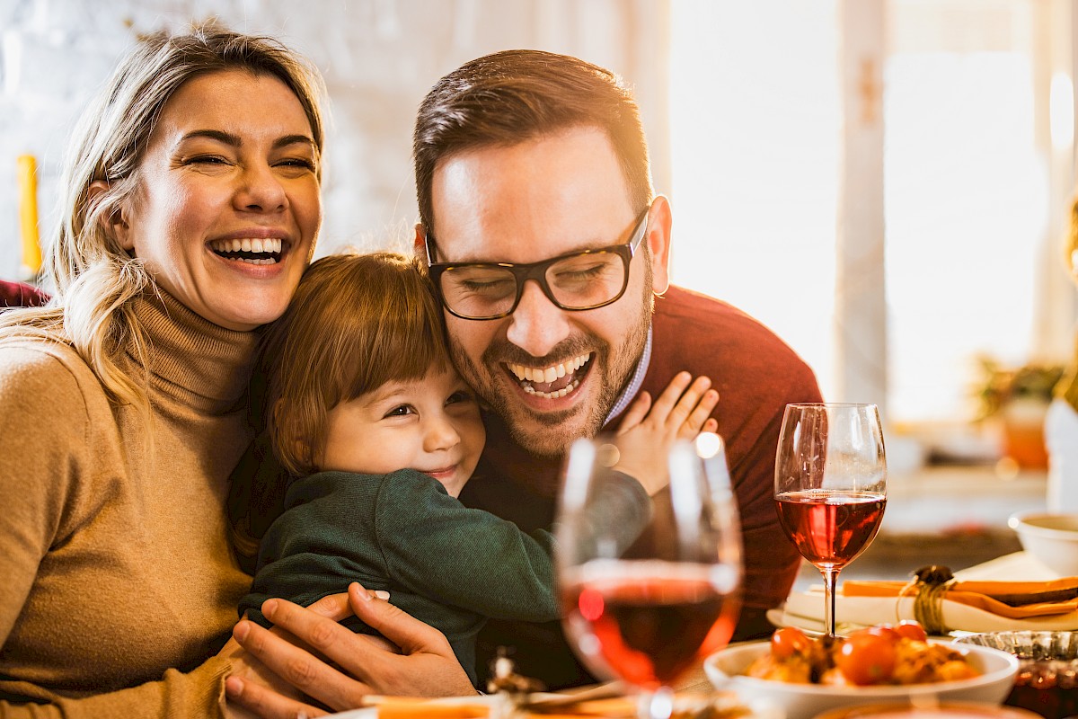 A happy family hugging and laughing together at a dinner table with wine and food in a cozy, warmly lit home.