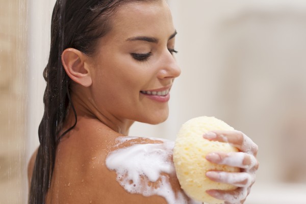A woman taking a shower, holding a sponge, with soap suds on her shoulder in a bathroom.