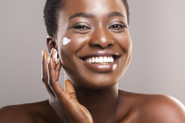 A smiling woman with short curly hair applies cream to her face, showcasing skincare and beauty routine.