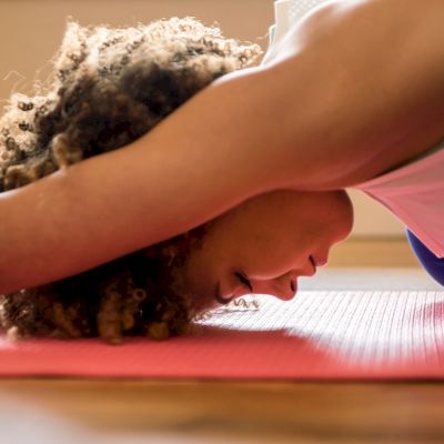 A person is practicing a yoga pose on a red yoga mat, stretching their arms forward with their forehead resting on the mat, indoors.