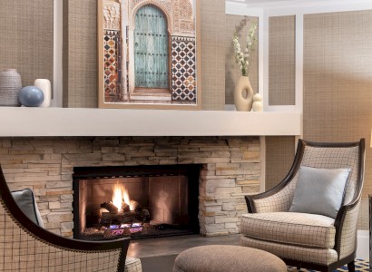 A cozy living room features a stone fireplace, two armchairs, a round ottoman, and artwork above, creating a warm, inviting ambiance.