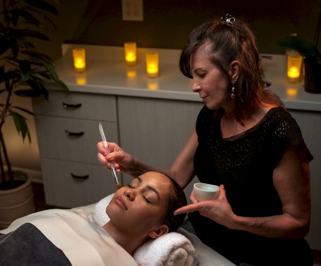 A woman is gently applying a facial treatment to another person lying on a bed in a dimly lit, candle-lit spa room.