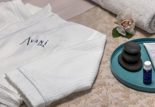 Spa setup with a white robe, orchid, rolled towel, skincare products, and massage stones on a decorative mat.
