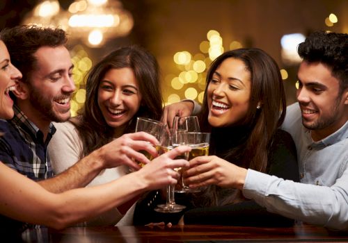 A group of five people is smiling and clinking their drinks together in a festive, warmly lit setting, likely celebrating or socializing.
