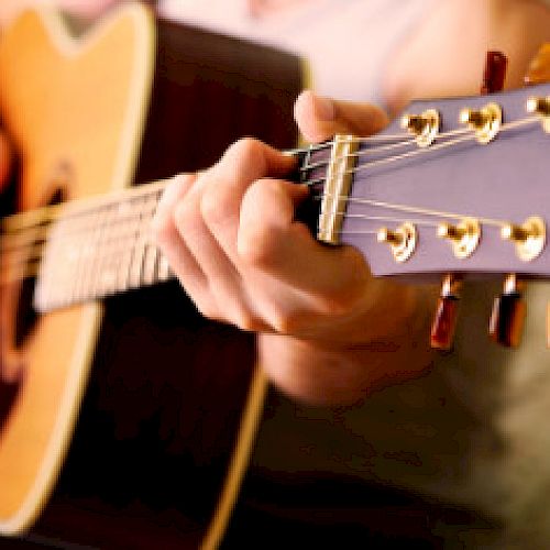 A person is playing an acoustic guitar, focusing on the fretboard and strings. The image captures the hand positioning and details of the instrument.