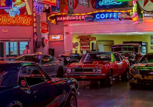This image shows a collection of classic cars displayed indoors, surrounded by vintage neon signs for various businesses like gas stations and diners.
