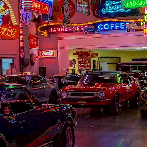 This image shows a collection of classic cars displayed indoors, surrounded by vintage neon signs for various businesses like gas stations and diners.