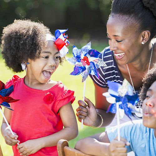 A woman and two children are enjoying a sunny day outside, playing with colorful pinwheels and smiling brightly.
