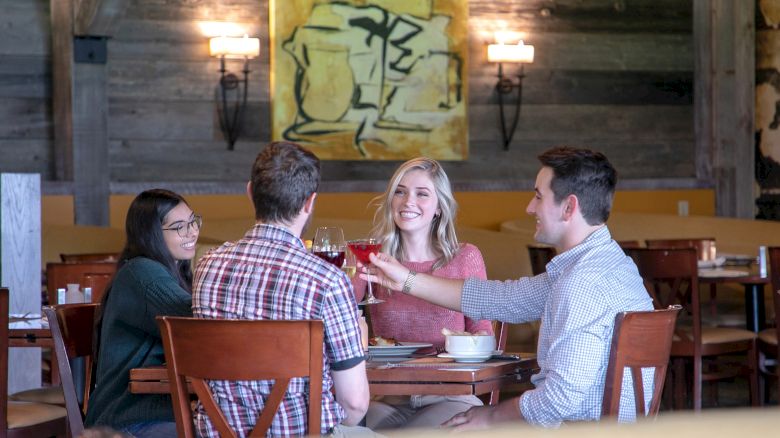 Four people are sitting at a restaurant table, smiling and toasting with drinks. There is abstract artwork on the wall behind them.