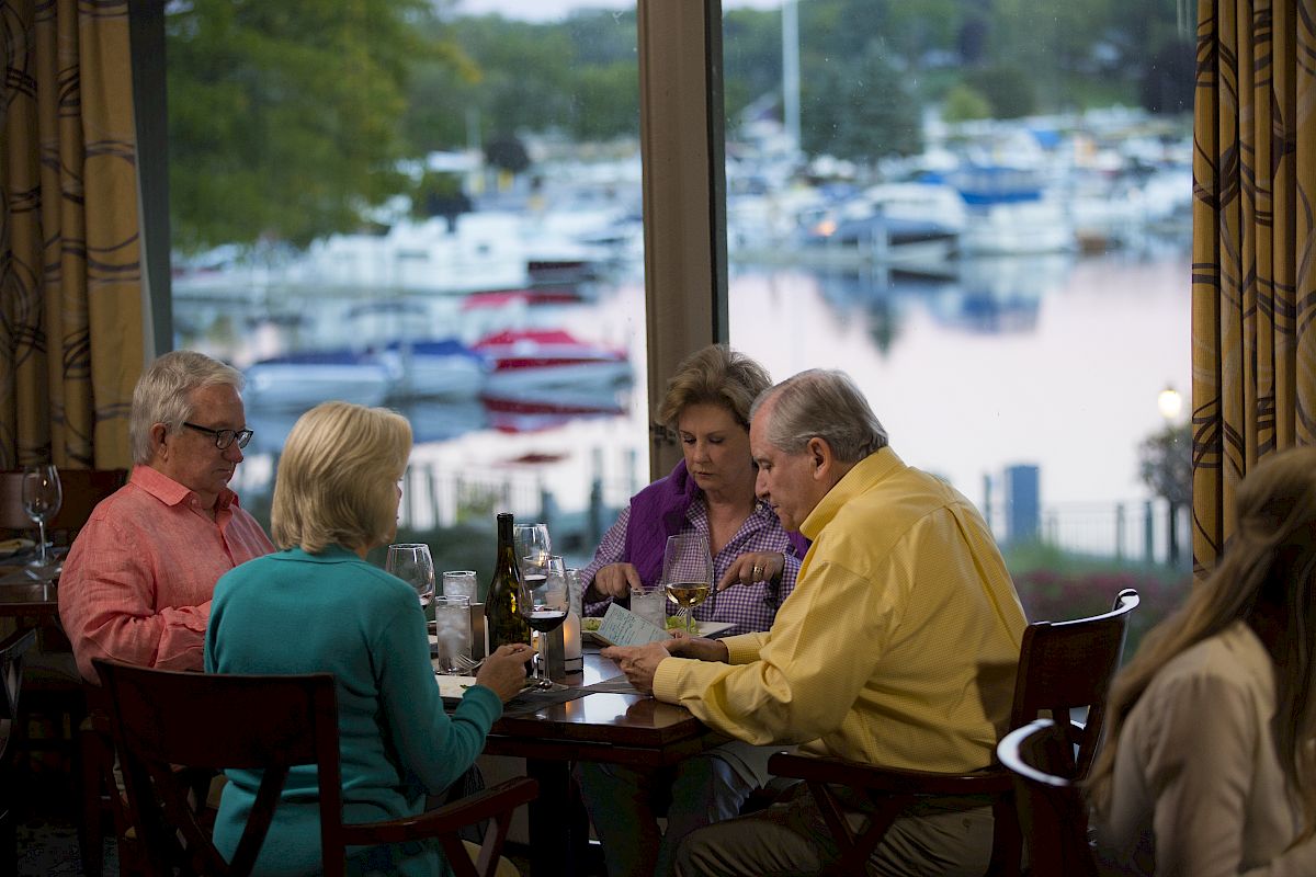 Four people are dining at a restaurant with a view of a marina full of boats seen through large windows, engaging in conversation.