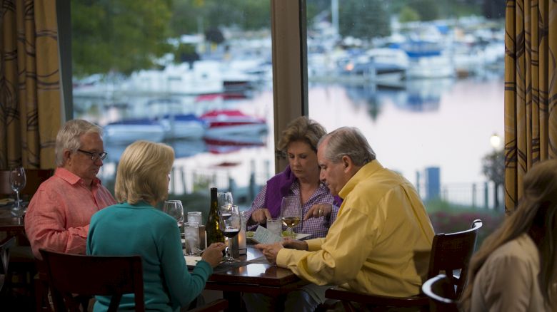 Four people are dining at a restaurant with a view of a marina full of boats seen through large windows, engaging in conversation.