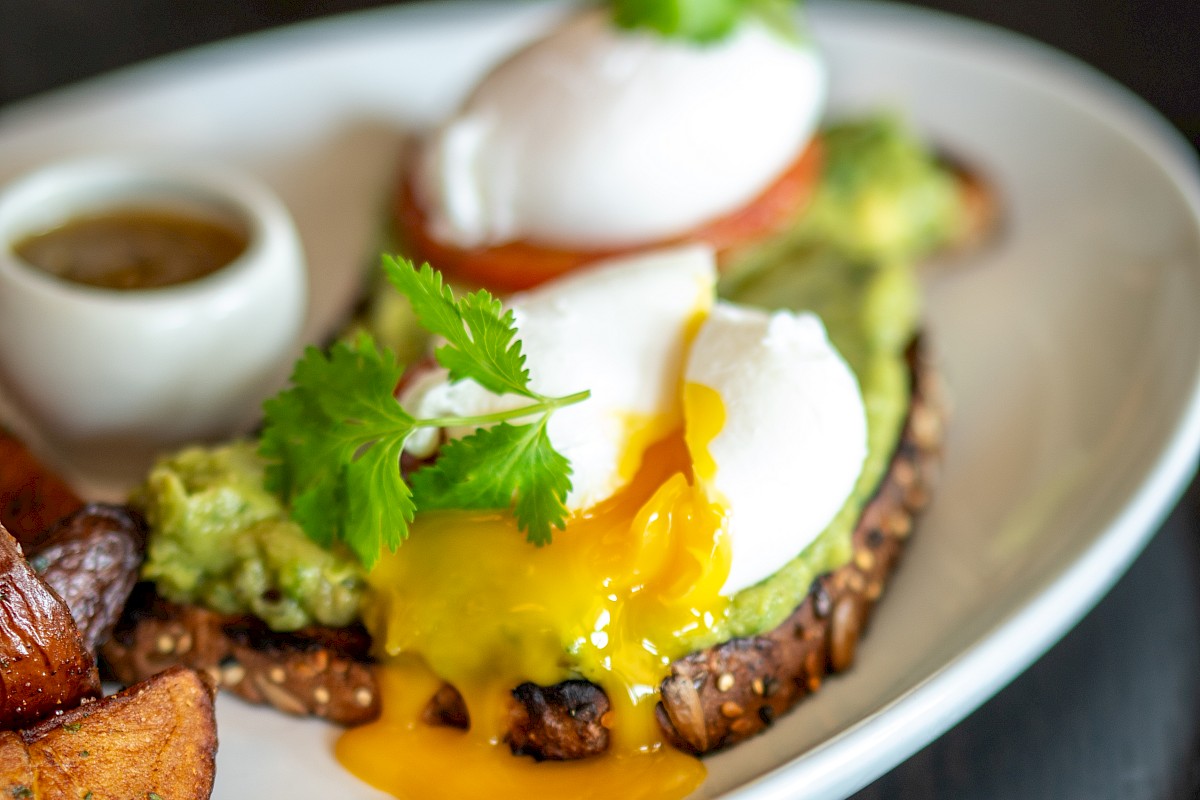 An open-faced sandwich with poached eggs, avocado, and garnish on toast, with roasted potatoes and a small sauce cup on the side.