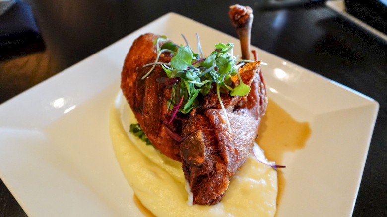 A roasted chicken leg served on creamy mashed potatoes, garnished with microgreens on a square white plate.