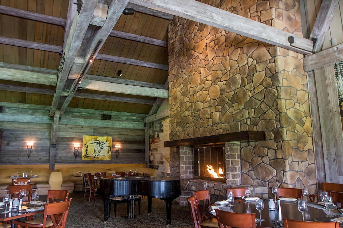 The image shows a cozy restaurant with wooden beams, stone walls, a large fireplace, a piano, and elegantly set tables and chairs, creating a warm ambiance.