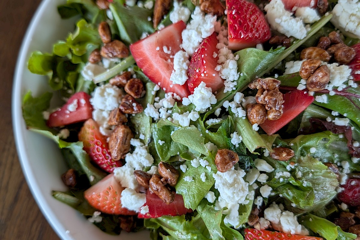 A fresh mixed salad with strawberries, crumbled cheese, nuts, and greens in a white bowl on a wooden table.