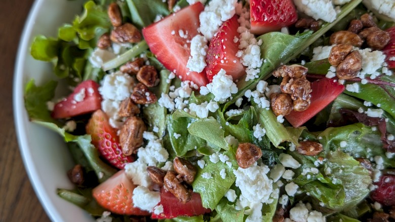 A fresh mixed salad with strawberries, crumbled cheese, nuts, and greens in a white bowl on a wooden table.