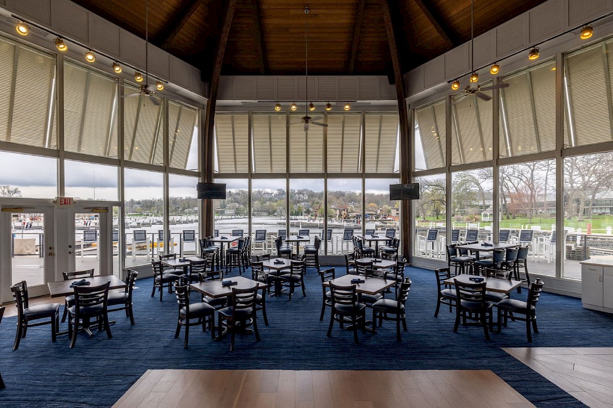 A spacious restaurant with large windows overlooking a waterway, featuring wooden beams, ceiling fans, and neatly arranged tables and chairs.
