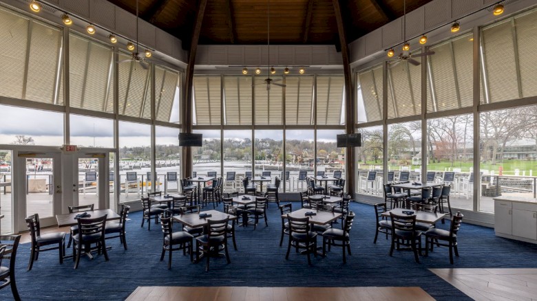 A spacious restaurant with large windows overlooking a waterway, featuring wooden beams, ceiling fans, and neatly arranged tables and chairs.