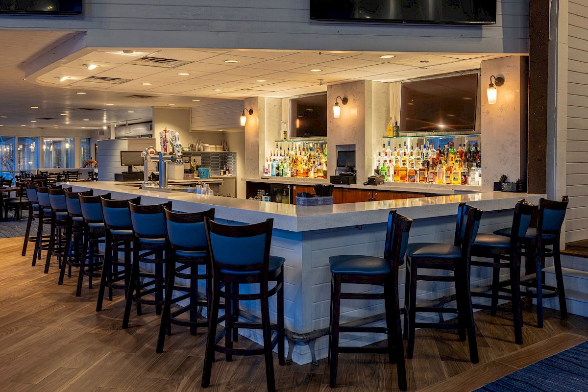 A well-lit bar area with a wide counter, black chairs, liquor bottles, and TV screens in a contemporary restaurant setting.