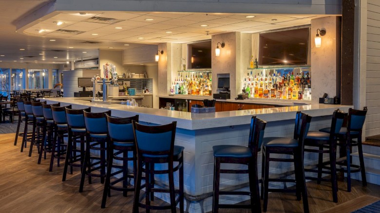 A well-lit bar area with a wide counter, black chairs, liquor bottles, and TV screens in a contemporary restaurant setting.
