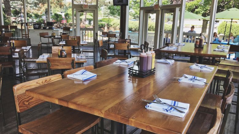 A restaurant with wooden tables and chairs, set with napkins and cutlery, and featuring large windows with a view of an outdoor seating area.