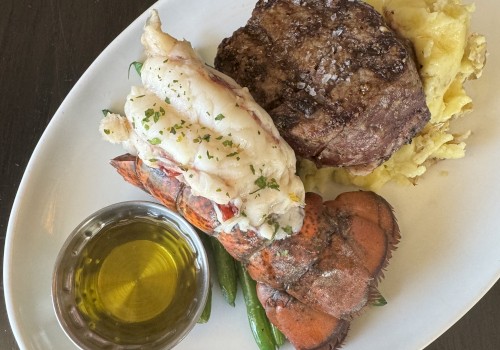 A meal with steak, mashed potatoes, lobster tail, green beans, and a small bowl of melted butter on an oval plate set on a dark table.