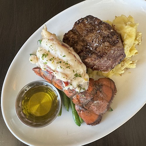 A meal with steak, mashed potatoes, lobster tail, green beans, and a small bowl of melted butter on an oval plate set on a dark table.
