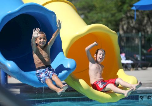 Two children are sliding down water slides into a pool, enjoying a sunny day.