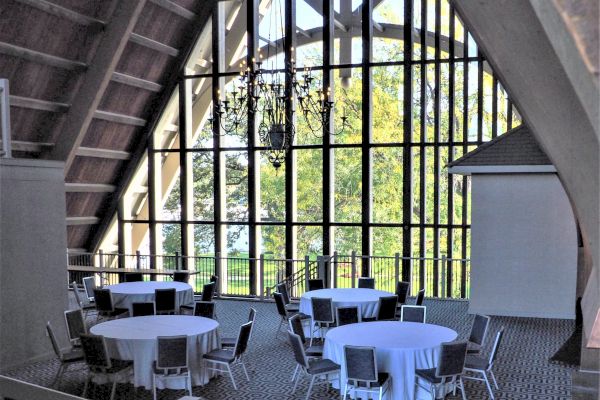 This image shows a room with large windows, round tables covered with white cloths, and chairs arranged around them, under a high ceiling.