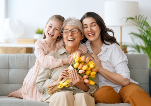 Three generations of women are smiling on a couch, with the grandmother holding a bouquet of yellow tulips.