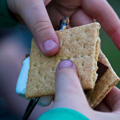 Two hands are seen making a s'more with graham crackers, chocolate, and a roasted marshmallow.
