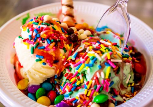 A bowl of colorful ice cream topped with sprinkles, candies, and a chocolate stick, with a plastic spoon on the side.