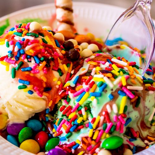 A bowl of colorful ice cream topped with sprinkles, candies, and a chocolate stick, with a plastic spoon on the side.