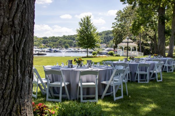 Outdoor setup with round tables and chairs, overlooking a lake with boats, surrounded by trees and a gazebo in the background.