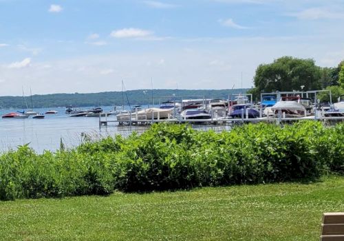 A lake scene with boats docked at a marina, lush green grass, and a bench in the foreground, under a partly cloudy sky ending the sentence.