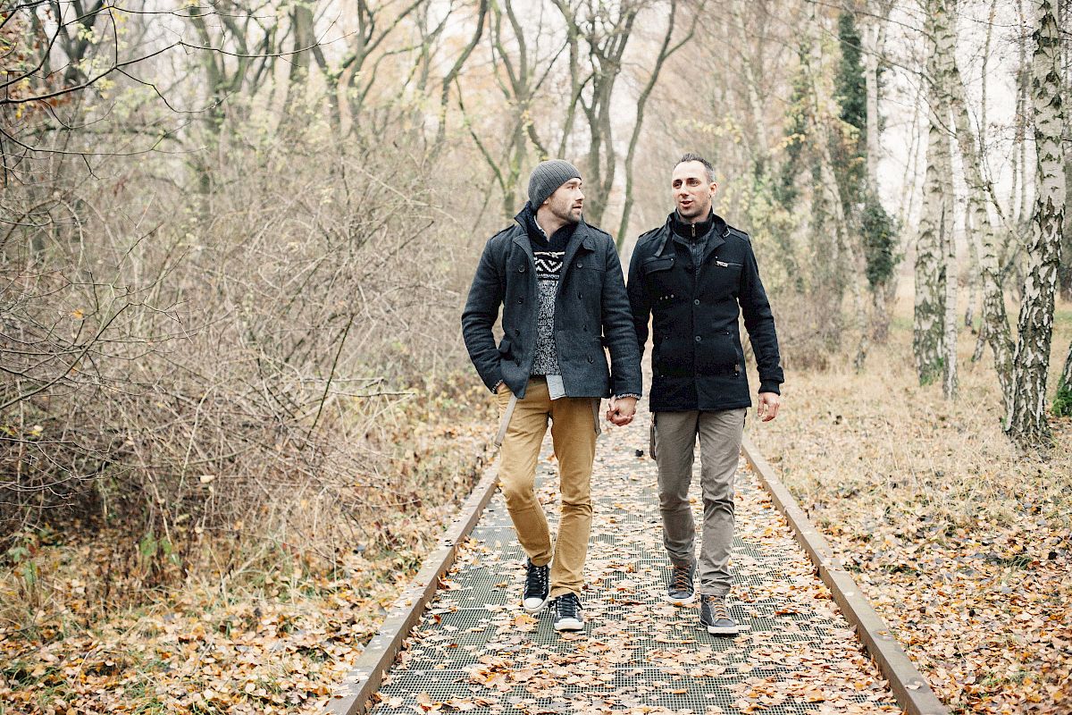 Two people walking on a leaf-covered path in a forested area, wearing jackets and holding hands.