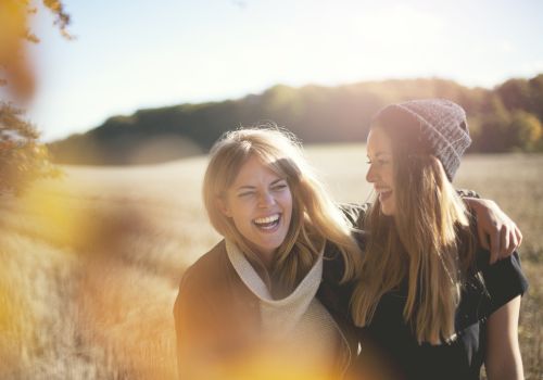 Two women are outdoors in a field, smiling and laughing with their arms around each other, enjoying a sunny day in nature.
