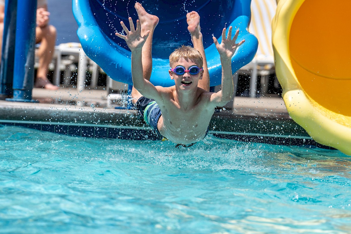 A boy with goggles is sliding out from a water slide into a pool, smiling and enjoying a sunny day at the water park.
