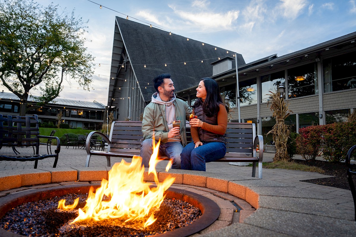 A man and woman sit on a bench around a fire pit, sharing drinks and smiling at each other during an outdoor evening.