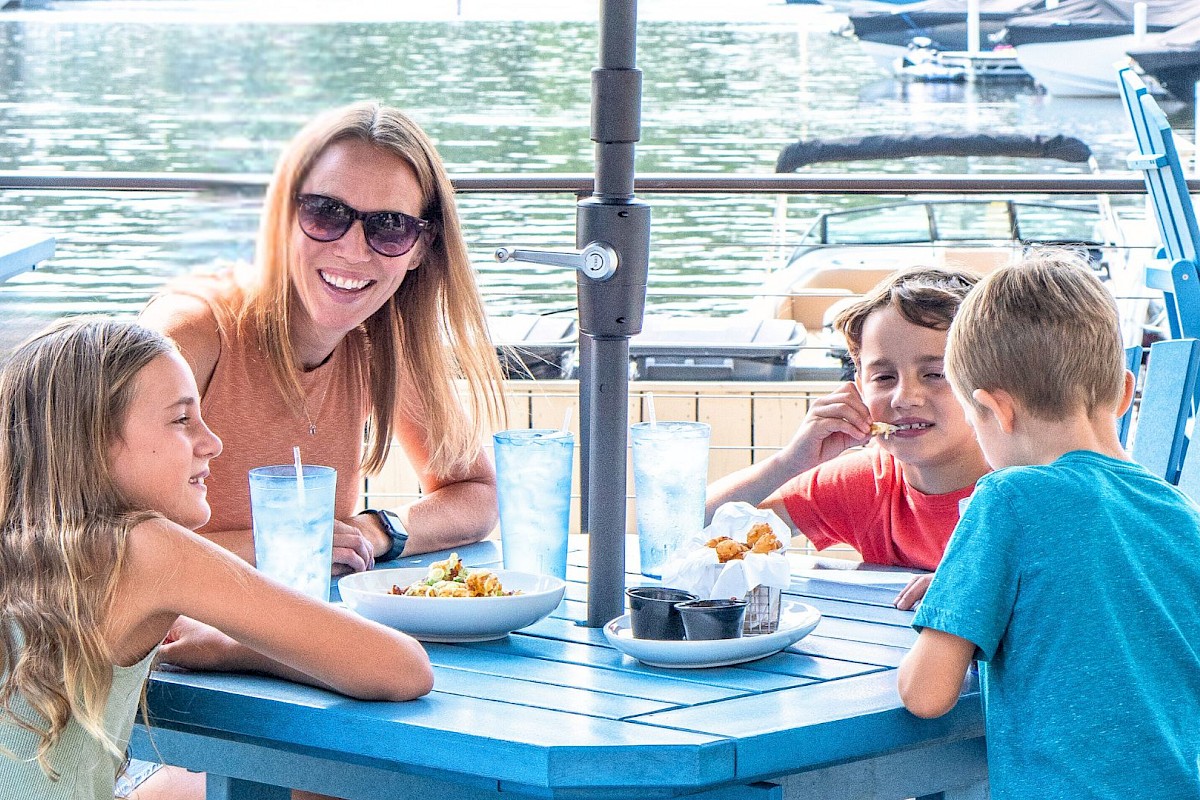 A woman and three kids are enjoying a meal outside by the water at a blue table with boats in the background.
