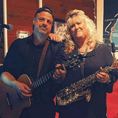 Two people pose with musical instruments, one holding a guitar and the other a saxophone, smiling indoors near a lit sign and wooden background.