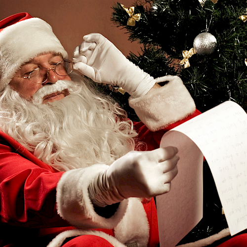 Santa Claus is sitting by a Christmas tree, wearing glasses and reading a long list.