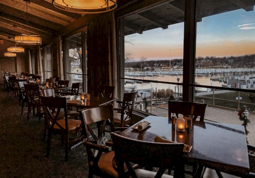 A chic, dimly lit restaurant interior with wooden chairs and tables, large windows overlooking a marina at sunset. End.