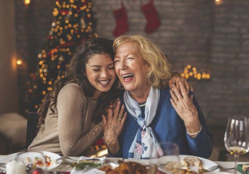 Two women share a joyful moment at a festive dinner table, with a decorated Christmas tree and stockings in the background.